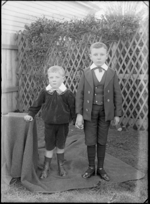 Two unidentified young boys outdoors standing on a blanket, shows tall wooden fence and shrubs behind them, probably Christchurch district