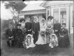 Wedding party portrait on grass in front of wooden house with veranda, unidentified bride with long veil and groom with extended family and flower girls [twins?], probably Christchurch region