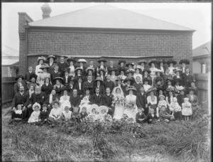 Large wedding group portrait on long grass in backyard with brick wall behind, unidentified bride and groom with extended family, children in front, women in hats, probably Christchurch region