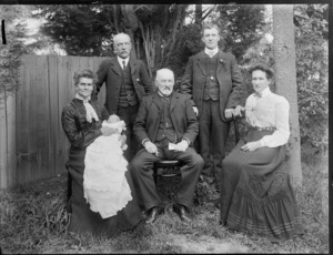Family portrait on long grass with wooden fence behind, unidentified men and women, one with lace collar and young baby in christening gown, probably Christchurch region