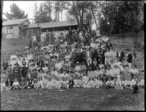 Large unidentified group of men and women, in hats on grass bank, children in front, with horse stables and tall trees behind, probably Christchurch district