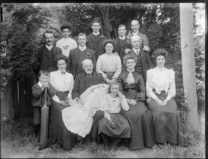 Portrait of extended family with young baby in christening gown, on long grass with wooden fence behind, probably Christchurch region