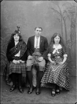 Portrait of an unidentified [performance?] group, showig two women and a man, with a painted backdrop, one woman wearing Scottish national costume including tartan skirt and sash and the other woman wearing a poker dot dress, embroidered with a harp, the man in knickerbockers, probably Christchurch district