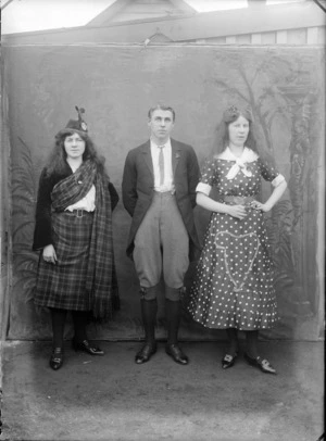 Portrait of an unidentified [performance?] group, showing two women and a man, with a painted backdrop, one woman wearing Scottish national costume including tartan skirt and sash and the other woman wearing a poker dot dress, embroidered with a harp, the man in knickerbockers, probably Christchurch district