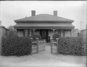 Unidentified elderly woman sitting on veranda of single story wooden house holding a cat with bird in cage nearby, with wooden gate and shrub fence in front, probably Christchurch region
