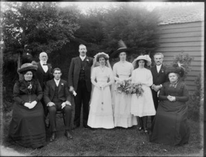 Wedding party portrait on grass next to wooden building with trees beyond, unidentified bride with long veil and groom with parents, best man and bridesmaids, women with hats, probably Christchurch region