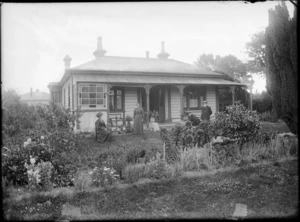 Exterior view of a wooden cottage, including a man and two women, all unidentified, standing on lawn in front, which is bordered by a garden, possibly Christchurch
