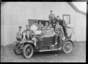 Group of unidentified men, sitting in and on a motorcar, which is parked outside a corrugated-iron [hall?] building, possibly Christchurch district