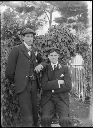 Two unidentified young men outdoors with a gate in the background, probably Christchurch district, both well dressed, wearing hats and flower corsages