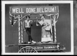 Two unidentified men dressed in costume as man and woman, standing on a wagon float with 'Christchurch Tramways' and 'Well Done, Belgium' signs, in front of wooden building, Christchurch
