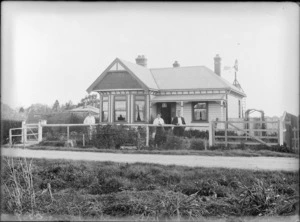 Exterior view of a single storied house, with a windmill mounted on roof, including unidentified women and a man, probably Christchurch district