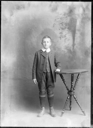 Studio portrait of unidentified boy wearing a suit and long woollen socks, standing next to a table, probably Christchurch district