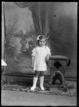 Studio unidentified family portrait of a young girl in a lace dress, curly hair, with white crochet socks and shoes with bows standing beside a wooden highchair, Christchurch