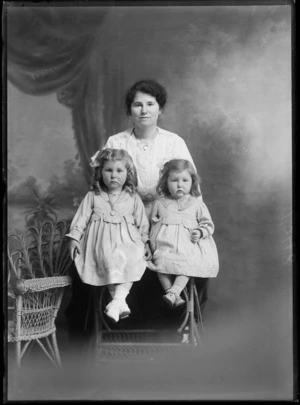 Studio unidentified family portrait of a young mother in a lace blouse and jacket, with heart shaped locket standing behind her two young daughters in matching dress, older with necklace, sitting on a cane table, Christchurch