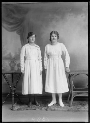 Studio portrait of two unidentified young women in white flower patterned dresses with belts standing between two tables, Christchurch