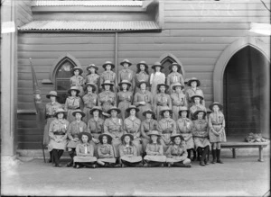 Group portrait of Sydenham Troop of the Girl Peace Scouts in front of church wooden wall, thirty young women and den mother in scout uniforms and hats, holding flag, Christchurch
