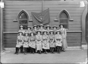 Group portrait of the Girl Peace Scouts, Senior Sydenham Troop, in front of church wooden sidewall, thirteen young women and den mother in scout uniforms and hats, with flag behind, Christchurch
