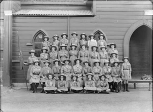 Group portrait of the Girl Peace Scouts, Sydenham Troop, in front of church wooden sidewall, thirty young women and den mother in scout uniforms and hats, holding flag, Christchurch