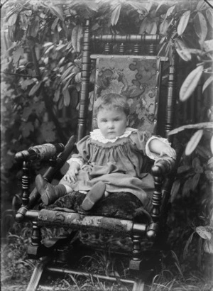 Portrait of young toddler in rocking chair in the garden in front of trees, with frilly lace neck and cuffs, probably Christchurch region