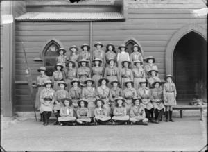 Group portrait of the Girl Peace Scouts, Sydenham Troop, in front of church wooden sidewall, thirty young women and den mother in scout uniforms and hats, holding flag, Christchurch