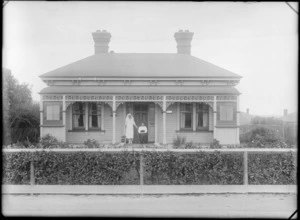 Exterior frontal view of a wooden cottage, including a sign [reading 'Wilson'?] next to front door, and iron fretwork around verandah, showing two unidentified women alongside, one possibly a nurse, [Christchurch district?]