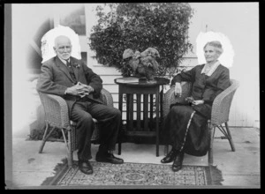 Unidentified elderly couple sitting on chairs, probably Hastings district, including pot plant, book and glasses on a table
