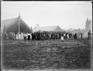 Women greeting vistors to gathering in front of meeting houses, Omahu Marae, with portraits and photos of family members, Fernhill, Hastings, Hawke's Bay District