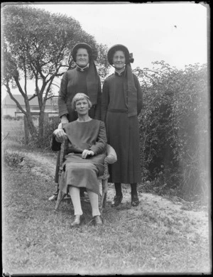 Group of women, two [Salvation Army?] women in uniform standing behind a seated woman in cane chair, garden lawn area, Hawke's Bay District