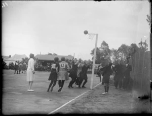North Island Netball Team on court in action, with spectators looking on, local school building and tent beyond, Hawke's Bay District