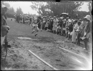 View of a primary school boy in mid-flight during the long jump competition, spectators and officials look on, Hawke's Bay District