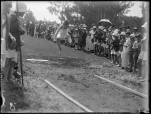 View of a primary school boy in mid-flight during the long jump competition, spectators and officials look on, Hawke's Bay District