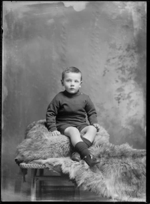 Studio unidentified family portrait of a young boy in a vest and shorts sitting on a fur rug covered cane table, Christchurch