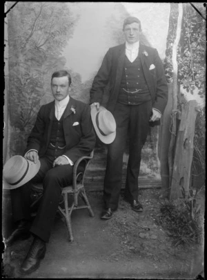 Unidentified wedding party portrait in the backyard in front of false backdrop next to wooden fence, best man standing with groom sitting in cane chair, with hats in hand, probably Christchurch region