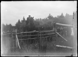 View of a horse and rider jumping a thicket fence with people looking on, trees beyond, location unknown