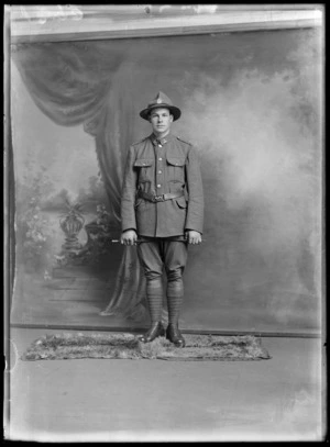 Studio portrait of unidentified young World War One soldier in uniform holding a swagger stick, with bird collar badges and hat with black band and silver fern crown badge, Christchurch