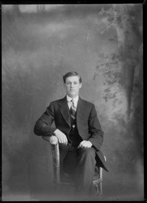 Studio portrait of unidentified young man with stiff arundel shirt collar, striped tie and greenstone pendant chest band, sitting in cane chair, Christchurch