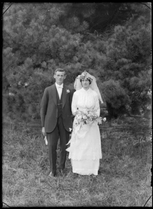 Outdoors wedding portrait, unidentified groom and bride with veil and holding flowers on long grass in front of large pine tree, probably Christchurch region