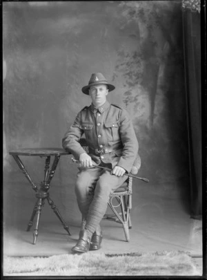 Studio portrait of unidentified young World War One soldier with hat and collar badges, stirrups and riding crop, sitting on cane chair, Christchurch
