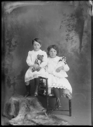Studio family portrait of unidentified young sisters in similar cotton lace dresses and pearl necklaces, holding either a doll or a teddy bear, sharing the same chair, Christchurch