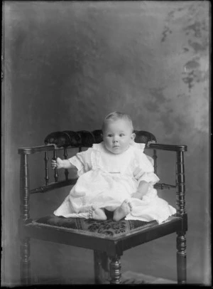 Studio portrait of an unidentified young baby in christening gown sitting on wooden high chair, Christchurch