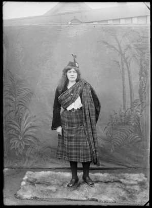 Outdoors portrait in front of false backdrop, an unidentified young woman in Scottish costume, tartan kilt and plaid sash with shoulder buckle, Glengarry bonnet with feather and badge with regimental insignia, probably Christchurch region