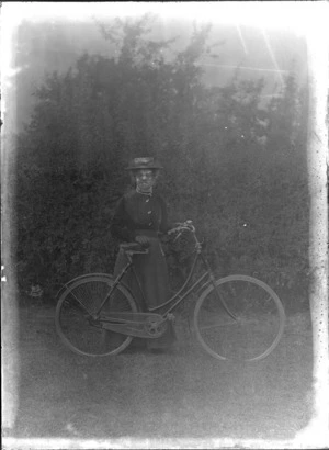 Unidentified woman on grass in front of a hedge, with a women's style bicycle, pearl necklace and straw hat, probably Christchurch region