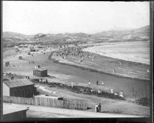 View over beach from road with buildings foreground, people on beach with changing sheds to distant hills with buildings and roads, [Lyall Bay, Wellington?]