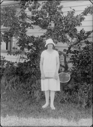 Outdoor portrait of unidentified young woman with hat and pale dress with pearl necklace, holding a tennis racket, probably Christchurch region