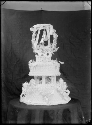 Two tiered wedding cake with figurines, horse shoes, flowers, pillars, wreath, bells, fern fronds, flowers and bow, probably Christchurch region