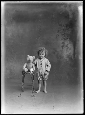 Studio portrait of an unidentified young child, wearing rompers and a shirt with a lace-up front, with a teddy bear, possibly Christchurch district