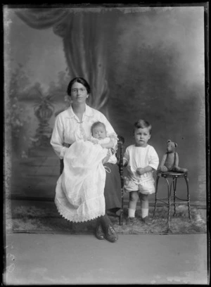 Studio portrait of an unidentified family group, showing a woman holding a baby wearing a [christening?] gown, and a small boy wearing rompers, with a teddy bear, possibly Christchurch district