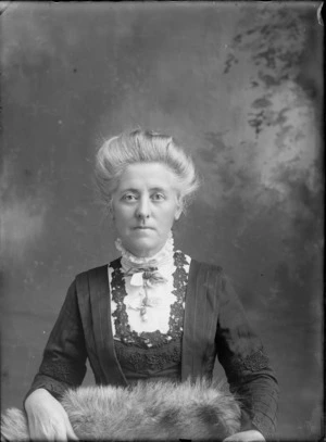 Studio upper torso portrait of an unidentified woman wearing a dark embroidered jacket over a lace high neck blouse, with a flower brooch at the neck, showing a fur rug across her lap, possibly Christchurch district