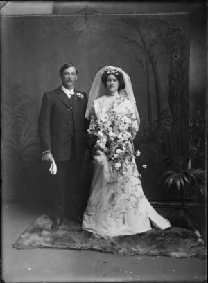 Studio wedding portrait of an unidentified couple, showing the bride and groom standing on a fur rug, with the bride holding a large bouquet of flowers, a floral arrangement in her hair attached to her veil, showing the groom with a posy in the button hole of his jacket, holding his gloves in his right hand, possibly Christchurch district