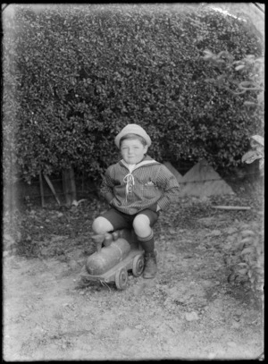 Outdoor portrait of an unidentified boy wearing a striped sailor shirt and hat, sitting on a wooden train engine, in a garden on an unidentified street, possibly Christchurch district